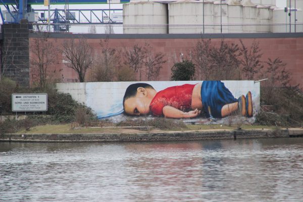 敘利亞庫德族男孩 Aylan Kurdi 在搭船逃難途中不幸溺斃，其遺體倒臥在海邊的照片迅速在全球流傳，引發國際社會對難民人道救援的關注。圖為出現在德國法蘭克福港口旁的塗鴉畫像。 圖｜Wikimedia